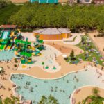 In this picture, we see the entire swimming area. There is a green lawn, various water slides, people swimming, and many towels on which people have placed their belongings.