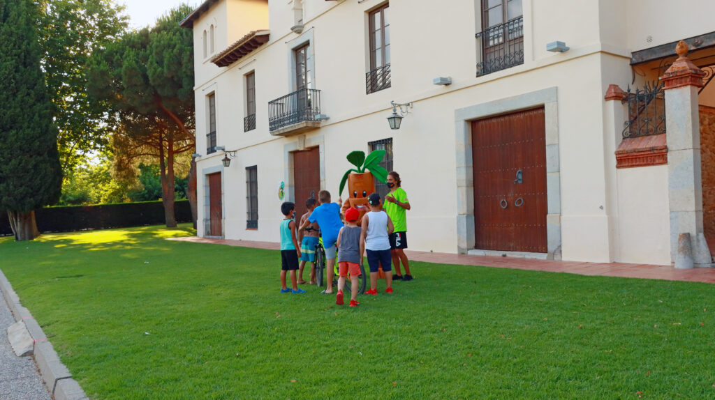 Children gathered around a mascot on the lawn in front of a white building